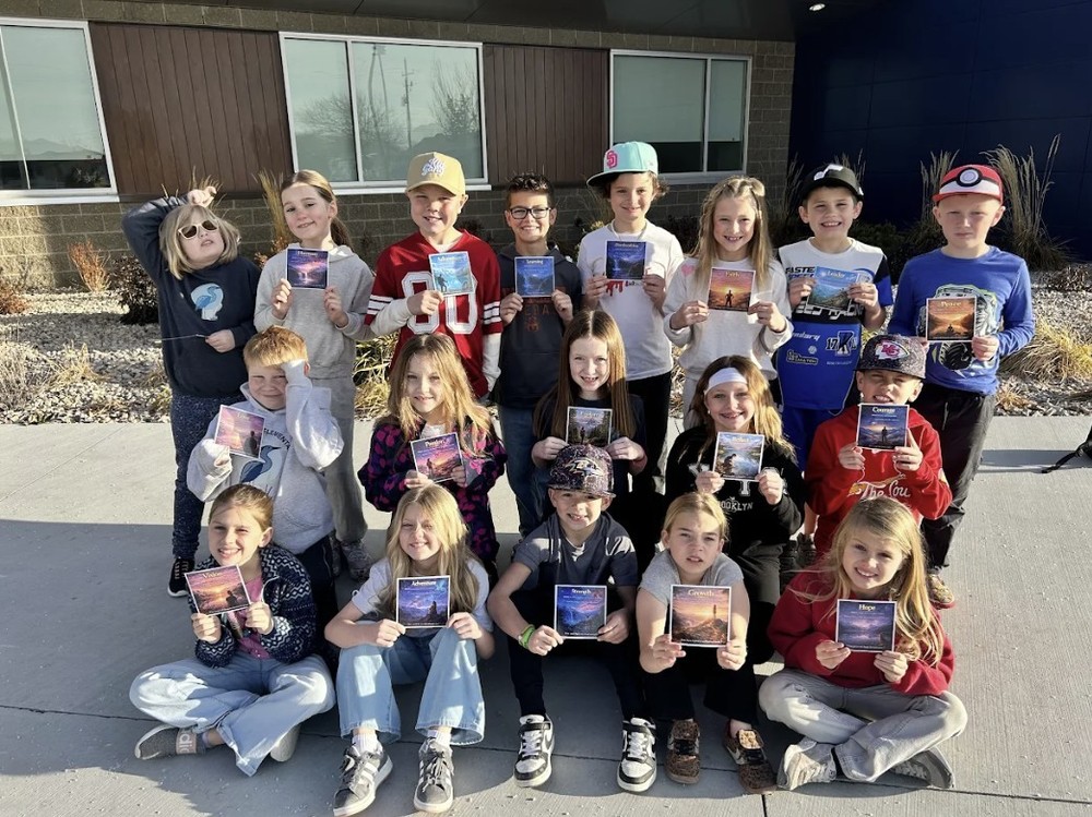 A group of sixteen elementary-aged children poses outdoors in front of a modern school building with wood and blue siding.