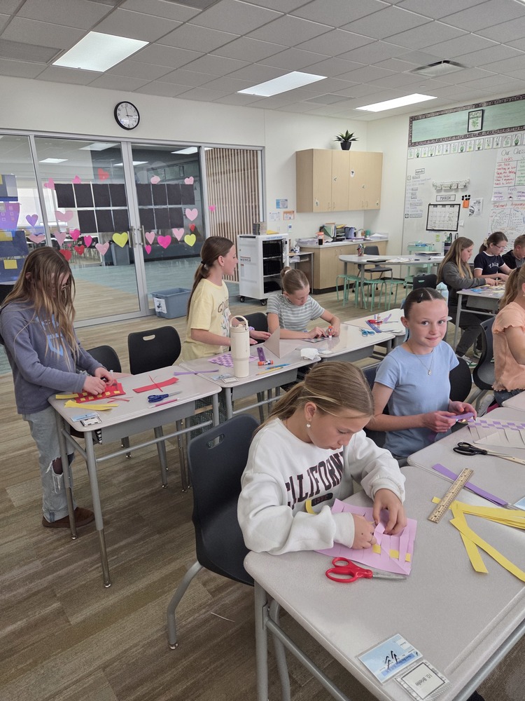 A brightly lit, modern classroom where several elementary-aged girls are seated at gray desks, focused on a paper-weaving art project. In the foreground, a girl in a white "CALIFORNIA" sweatshirt weaves yellow paper strips into a pink base. To her right, another girl in a light blue shirt smiles toward the camera. Other students are visible in the background using scissors and glue sticks. The room features a glass wall decorated with pink and red paper hearts, a white storage cart, and a large whiteboard filled with handwritten classroom schedules and "Math" charts.