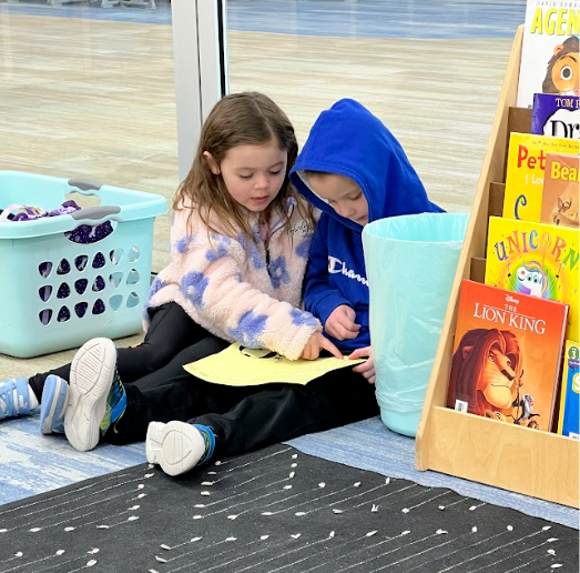 Two young children sit together on a blue carpeted floor next to a wooden book rack. A girl in a floral fleece jacket points to a yellow piece of paper held by a boy in a blue "Champion" hooded sweatshirt. In the background, a book rack displays titles including "The Lion King," "Unicorn," and "Pete the Cat."