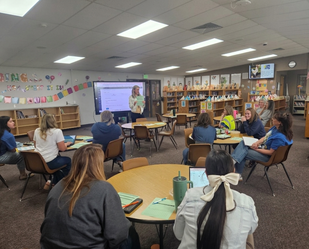 Lomond View Elementary teachers gathered around circular wooden tables in a modern school library. A facilitator in the middle distance is leading the group while holding a green worksheet and referring to a large presentation monitor displaying a spreadsheet. The environment is warm, with natural light and sharp details on materials like laptops, notebooks, and a green tumbler. The walls feature cheerful decorations and organized bookshelves.