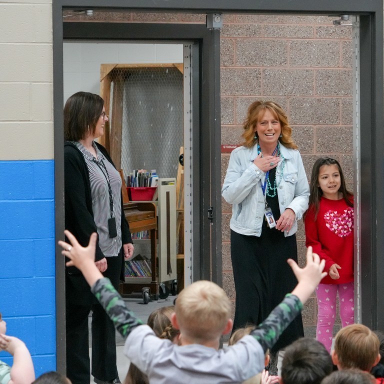 Principal Kari Breeding enters a room filled with cheering students, her hand over her heart in a gesture of surprise and gratitude.
