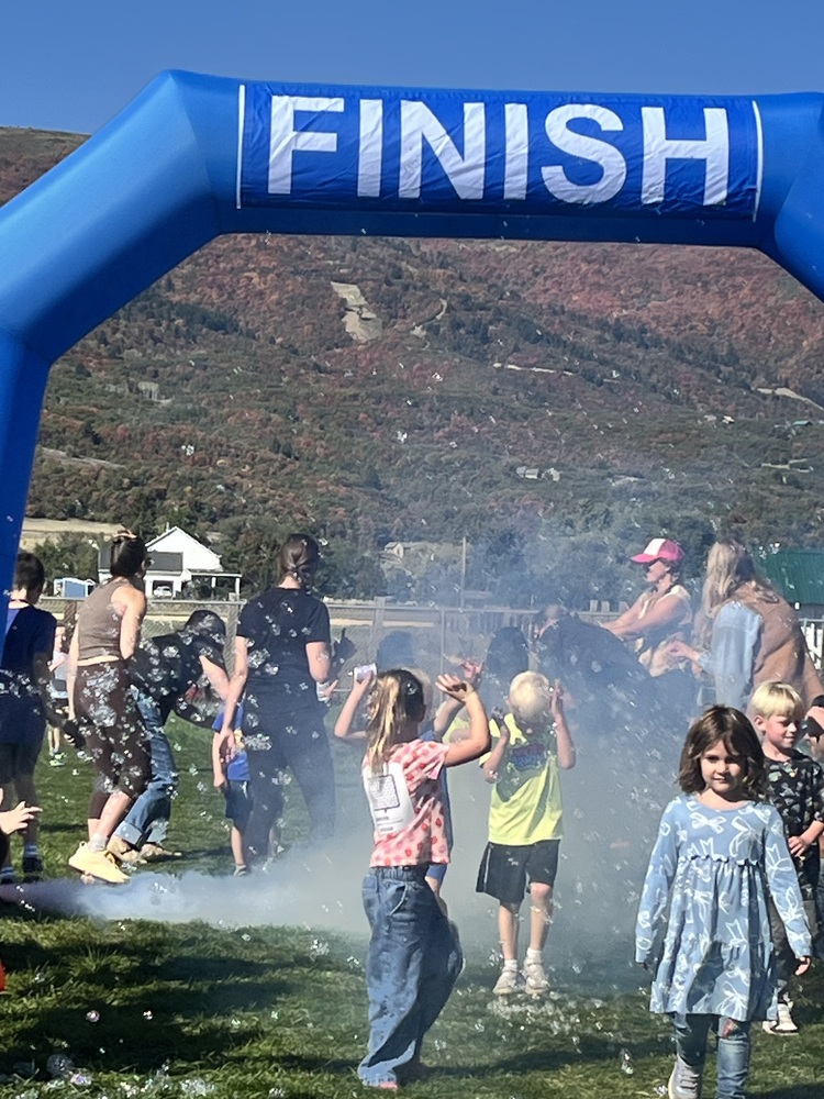 Children playing and running through bubbles and smoke beneath a large blue inflatable finish line arch during an outdoor event with mountains in the background.
