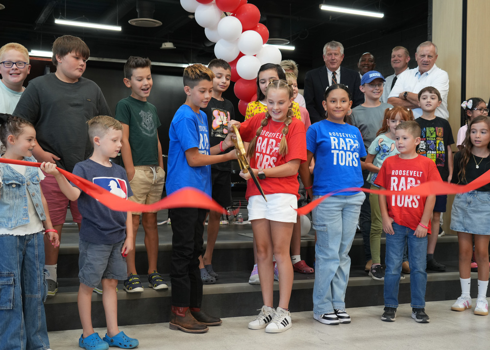 Students standing behind a red ribbon, two students with large scissors  cutting the ribbon