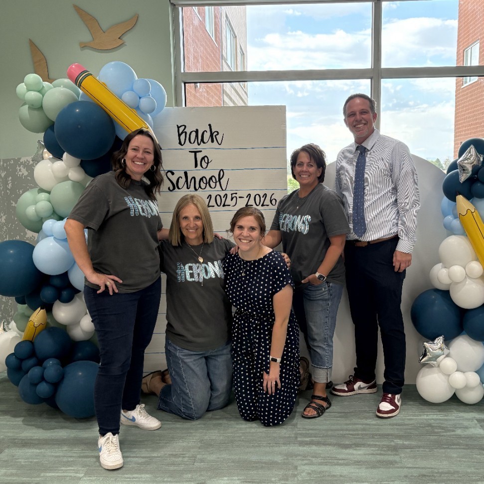 Five adults are standing and kneeling in front of a balloon arch decorated with pencils and stars. Behind them is a sign that looks like notebook paper with the words “Back To School 2025–2026.” Four of the adults wear matching gray shirts that say “HERONS.”