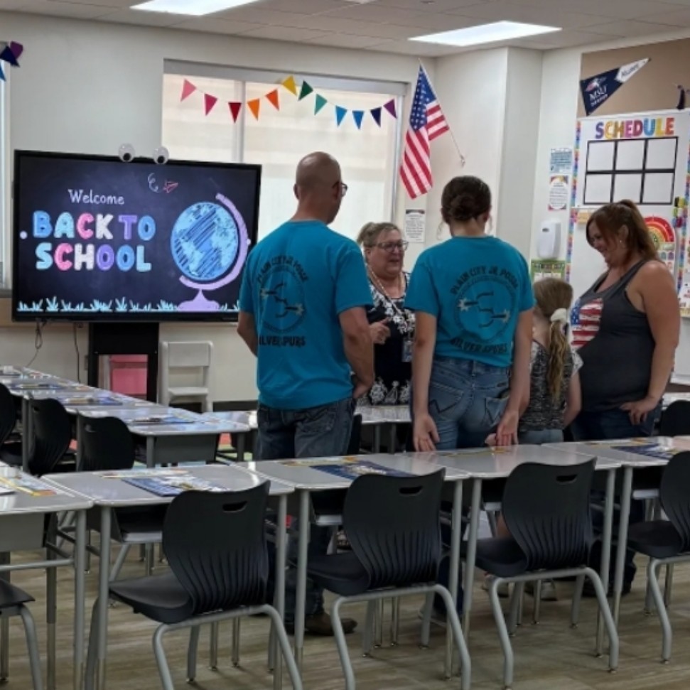 Ms. James' classroom set up with rows of desks and papers laid out. A group of adults and a child are gathered near the front talking to a teacher. On a large screen, colorful text reads “Welcome Back To School.” Bright triangular banners hang above the windows, and an American flag and a classroom schedule are visible on the wall.