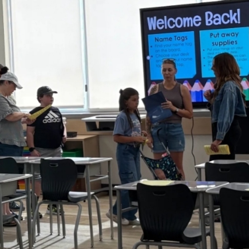 A classroom scene with parents and children lined up to speak with a teacher on the first day of school. A large screen in the background reads:
"Welcome Back!"
Name Tags: "Put your name tag on the board. Choose a desk and sit for name bingo."
Put away supplies: "Put supplies in your desk or cubby. Label items that aren't already."