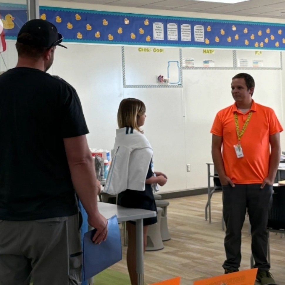 Mr. Weston, in an orange shirt, speaks with a student and her parent in a classroom doorway. Inside the classroom, a bulletin board titled “Class Goals” is visible, with sections labeled "Math", "ELA", and more. Rubber duck decorations line the board.
