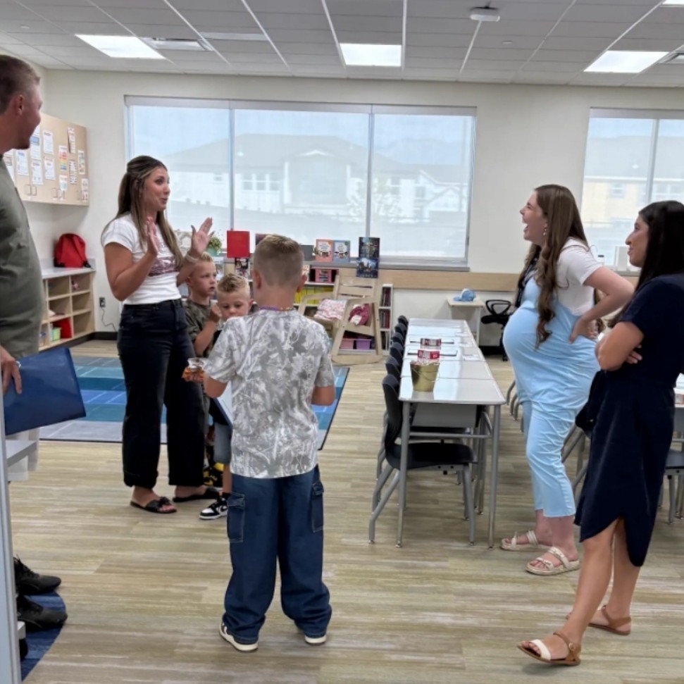 Mrs. Warr talks animatedly with a group of students and parents during an open house or meet-the-teacher event. Desks are organized in groups, and classroom materials are neatly arranged on shelves.