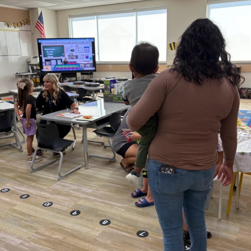 A woman holding a toddler enters a classroom where Mrs. Wilcox is seated at a table speaking to a young girl. A screen at the front displays a message:
"Welcome to our class!"
It includes visuals of the alphabet, numbers, and a rainbow-themed design. Classroom stations and colorful decor are visible throughout the room.