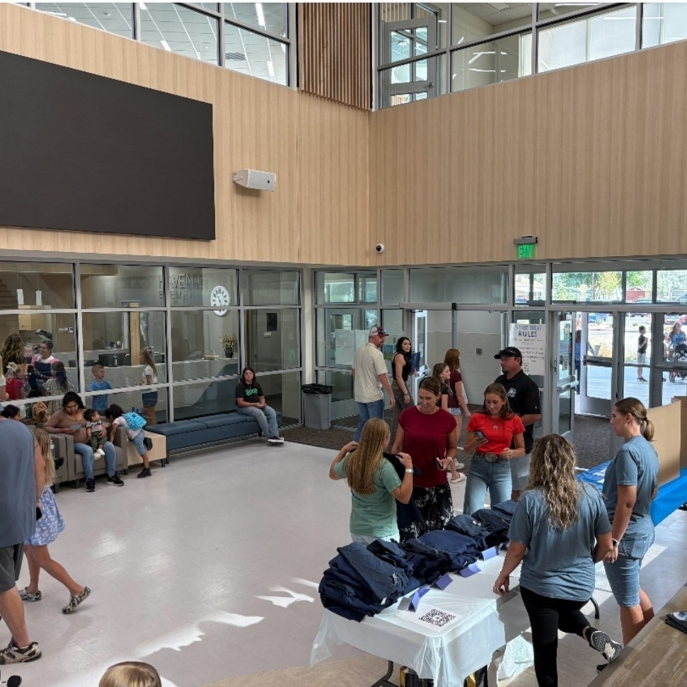 A busy school lobby or common area during a back-to-school or registration event. Several people are gathered at a table with folded navy blue shirts, where volunteers in blue shirts assist parents and students. On the table, a QR code and sign-up sheets are visible. In the background, families are seated on benches and walking through the entrance. A large, blank black display screen is mounted on a wood-paneled wall above.