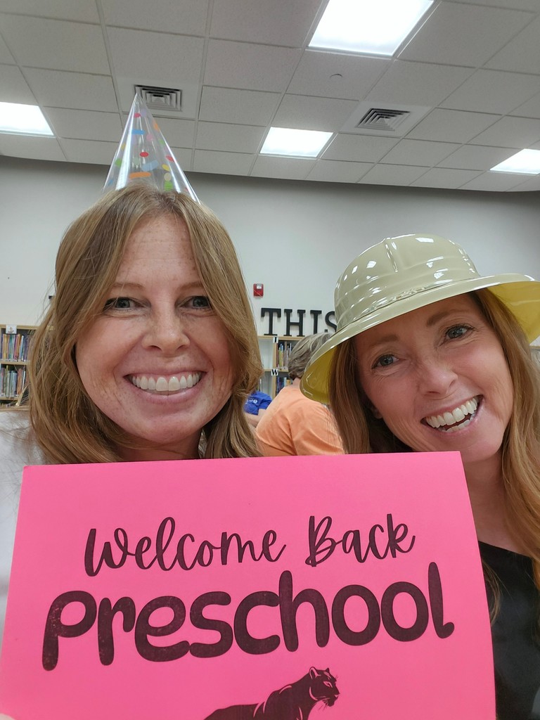 Two preschool teachers smiling and wearing silly hats holding a "welcome back preschool" sign