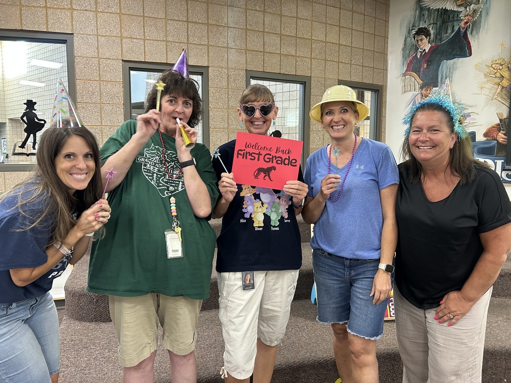 art teacher and first grade teachers holding a "welcome back first grade" sign and smiling with funny hats and magic wands and kazoos in their mouth