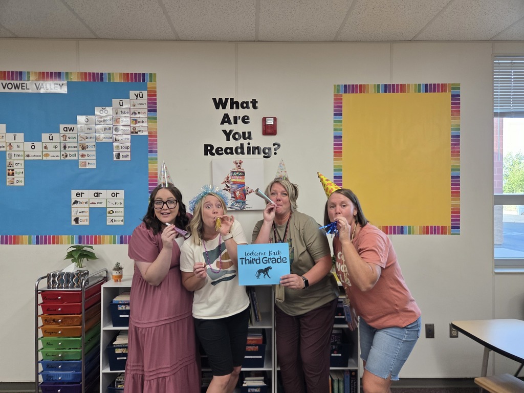 third grade team is making silly faces, blowing air whistles, wearing party hats, and holding a sign that says "welcome back third grade" there is a sign behind them that reads "what are you reading?" they are in a classroom 
