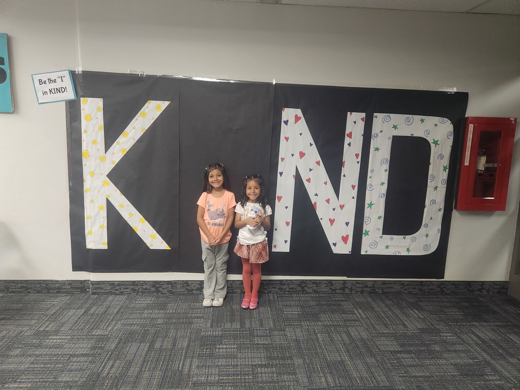 Students in front of black sign with bold papered letters spelling out the work KIND with students posing as the 'i' 