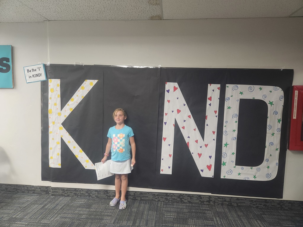 Students in front of black sign with bold papered letters spelling out the work KIND with students posing as the 'i' 