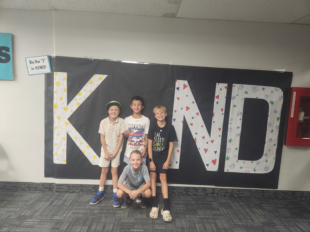 Students in front of black sign with bold papered letters spelling out the work KIND with students posing as the 'i' 