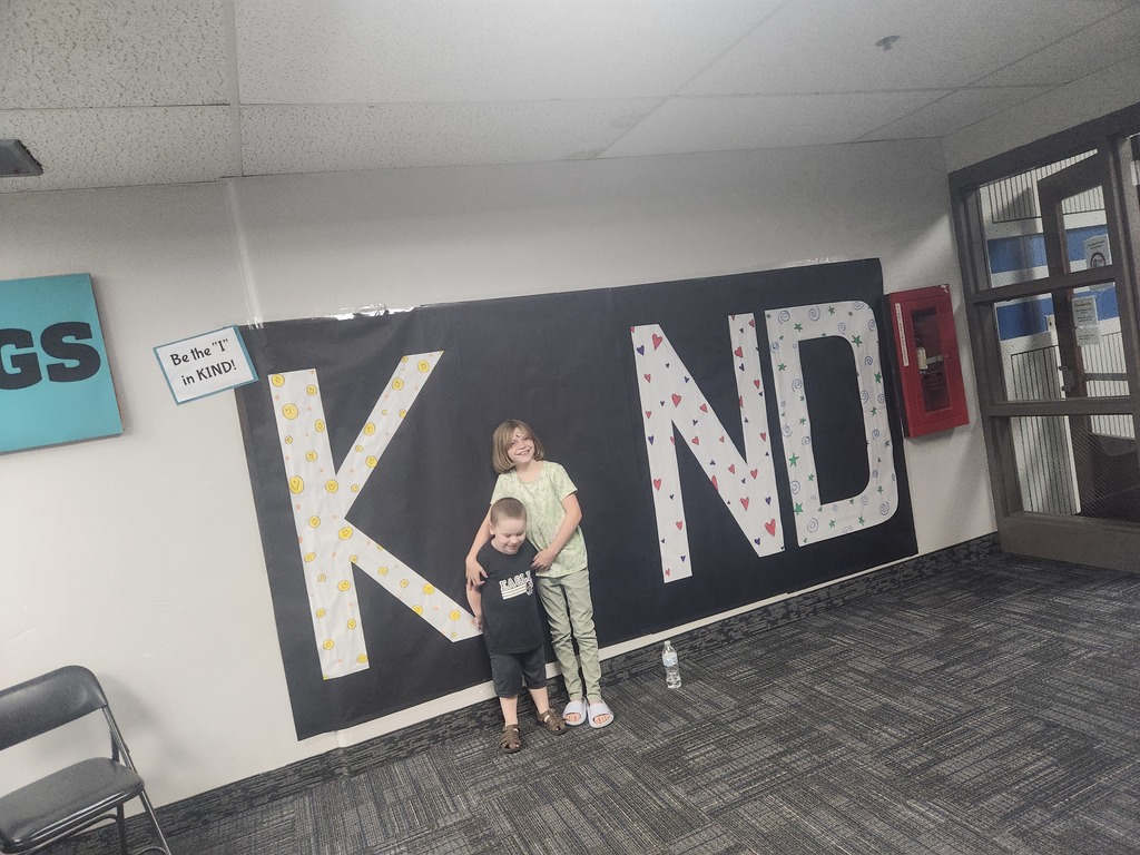 Students in front of black sign with bold papered letters spelling out the work KIND with students posing as the 'i' 