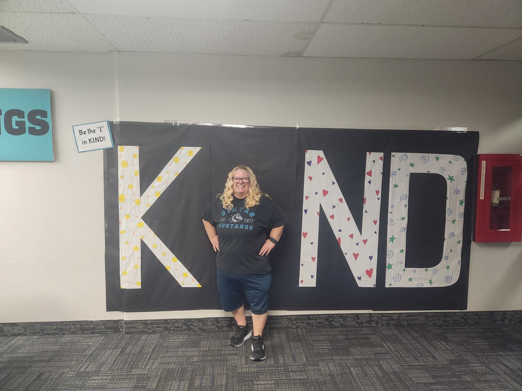 Mrs. Porter- 4th grade teacher- in front of black sign with bold papered letters spelling out the work KIND with Mrs. Porter posing as the 'i' 