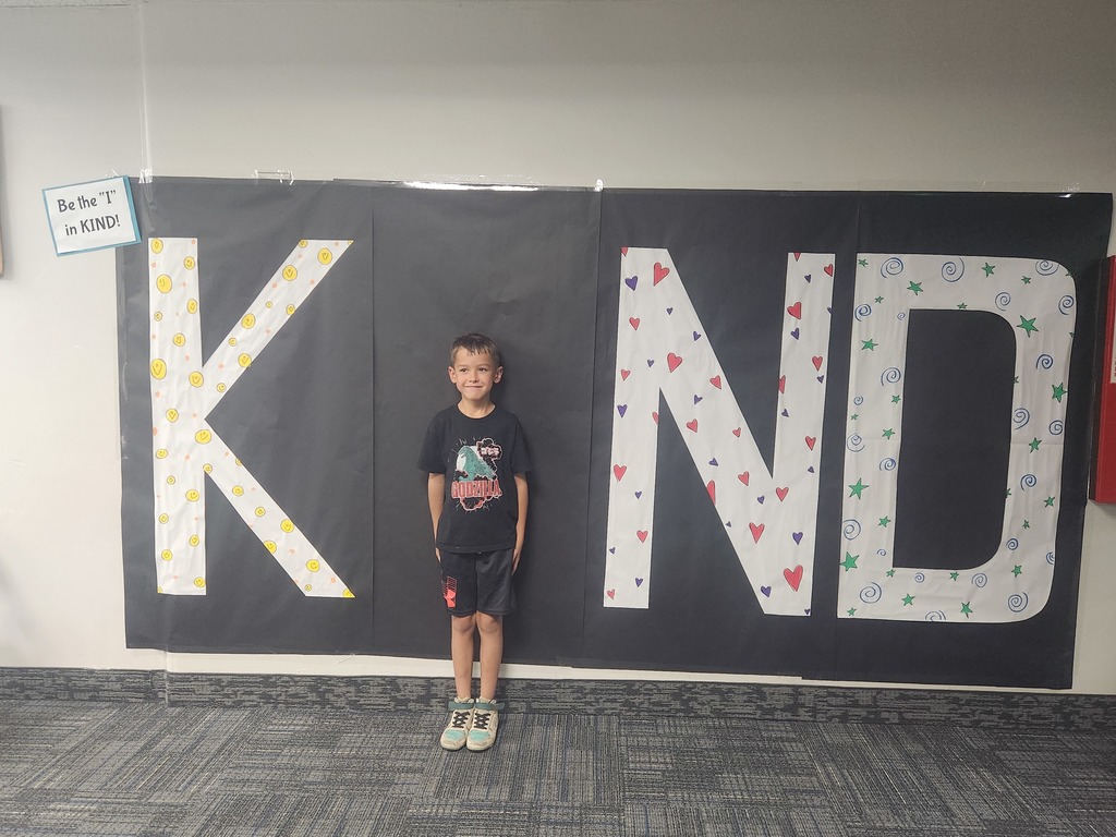 Students in front of black sign with bold papered letters spelling out the work KIND with students posing as the 'i' 