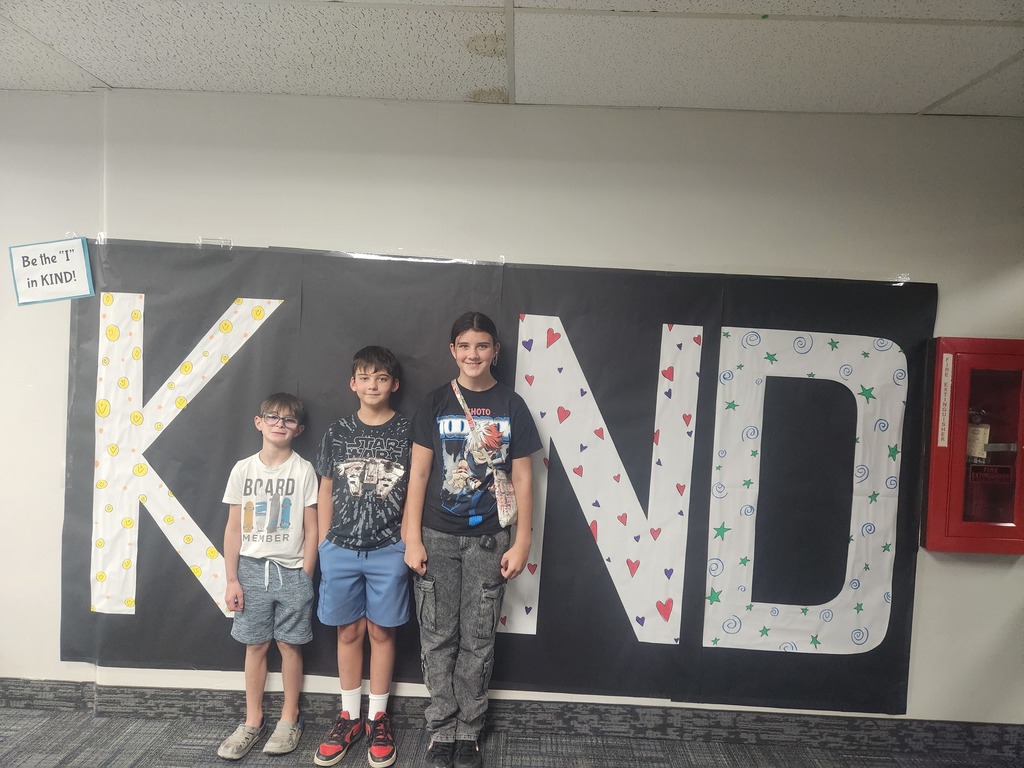 Students in front of black sign with bold papered letters spelling out the work KIND with students posing as the 'i' 