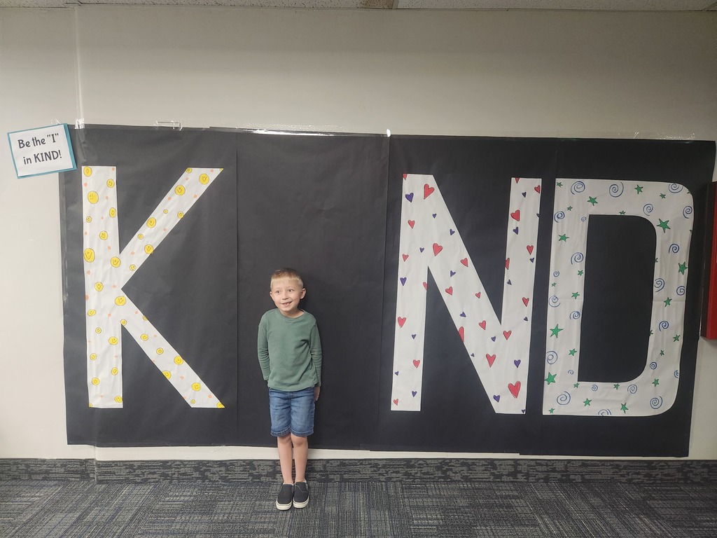 Students in front of black sign with bold papered letters spelling out the work KIND with students posing as the 'i' 