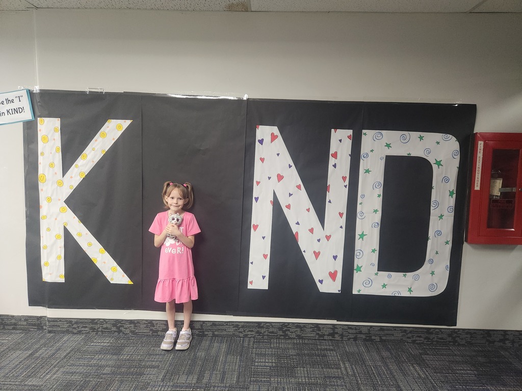 Students in front of black sign with bold papered letters spelling out the work KIND with students posing as the 'i' 