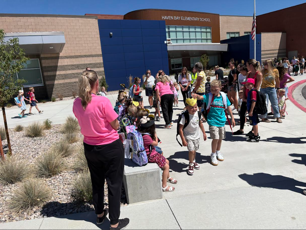 A group of elementary students and parents gathered outside Haven Bay Elementary School on a sunny day. Children are wearing backpacks, and staff members in bright pink shirts help guide the crowd.