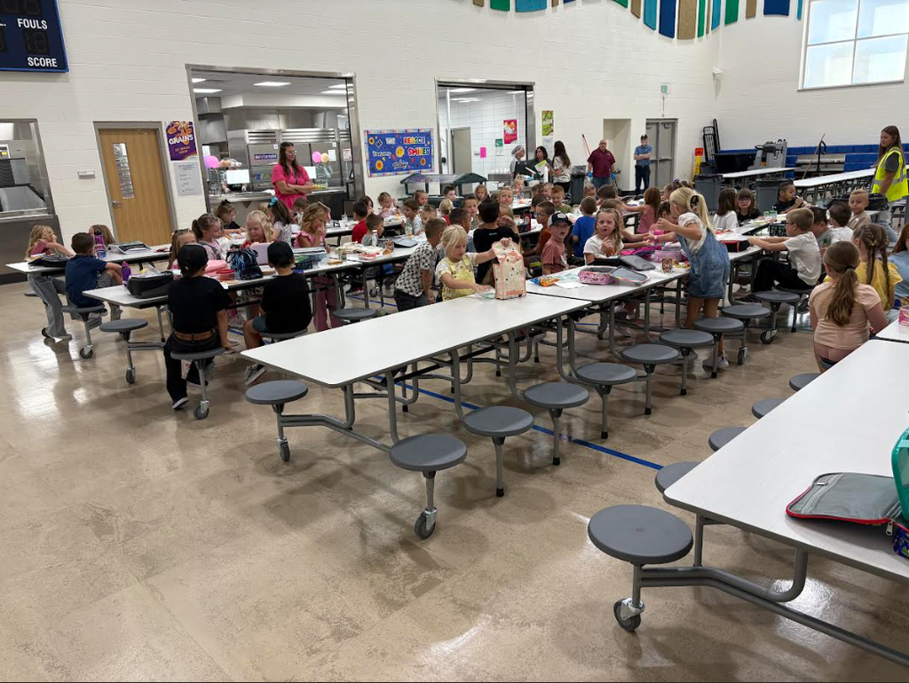 A lively cafeteria scene at an elementary school, with dozens of young students sitting at white tables eating lunch and chatting. Staff members are supervising in the background.