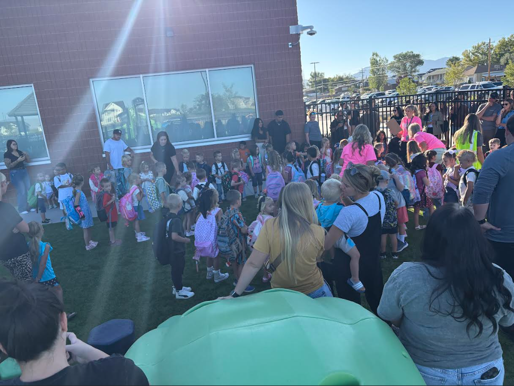 A large group of elementary students and their families gathered outside a school building for morning drop-off. Teachers and staff in pink shirts are helping organize the excited crowd of children wearing backpacks.