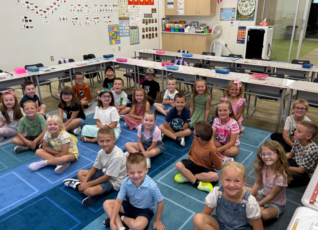 A large group of smiling elementary students sits on a blue classroom rug, facing the camera. Behind them are desks and a colorful classroom wall with charts and alphabet posters. The students are a mix of boys and girls in cheerful first-day outfits.