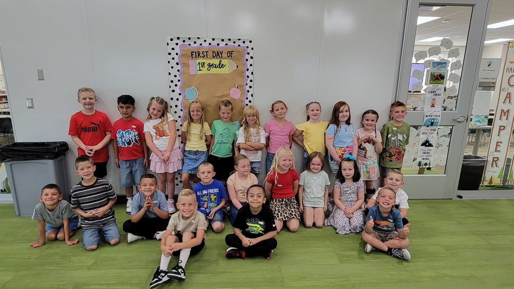 A class of first graders stands and kneels in front of a bulletin board that reads "First Day of 1st Grade." The students are smiling and grouped together, dressed in colorful clothes. The background includes classroom decorations and a door with encouraging signs.
