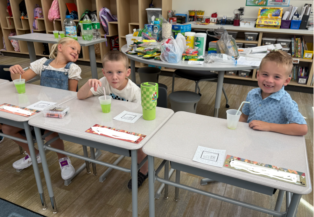 Three young elementary students sit at desks in a bright classroom, enjoying green drinks with straws. Two boys and one girl smile and interact happily. School supplies and backpacks are visible in the background.