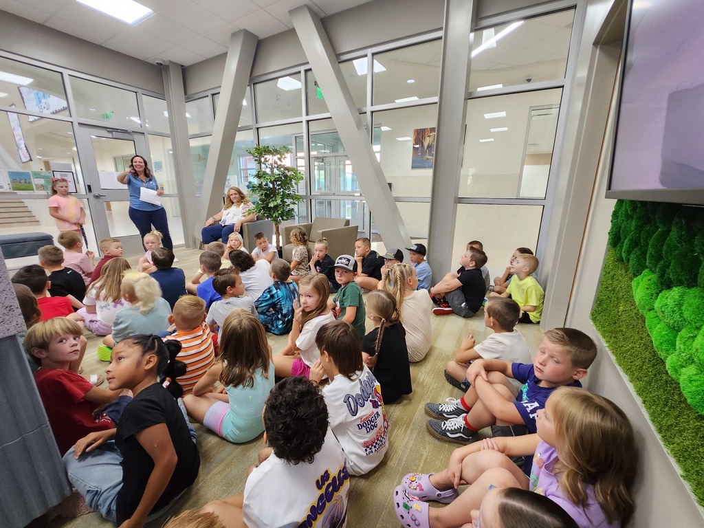 A large group of elementary students sits on the floor in a school office, attentively listening to two adults speaking. One adult stands with a paper in hand while the other sits in a chair. The office has glass walls and a modern design with natural light.