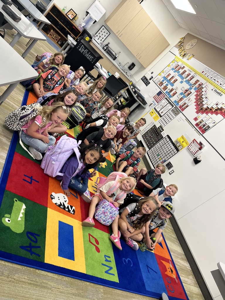 A group of young students sits on a vibrant classroom rug with letters and animals, posing for a photo. Many have backpacks and are smiling brightly. The classroom is decorated with learning charts and has a cozy, welcoming atmosphere.