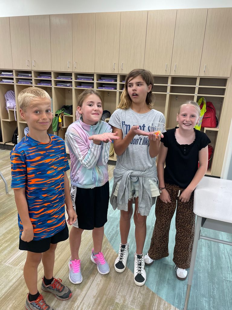 Four children stand in a classroom in front of cubbies, smiling and posing for the camera. One girl holds a small plastic container with candy corn. The students are casually dressed, and the classroom setting includes wooden storage cubbies with backpacks and books.