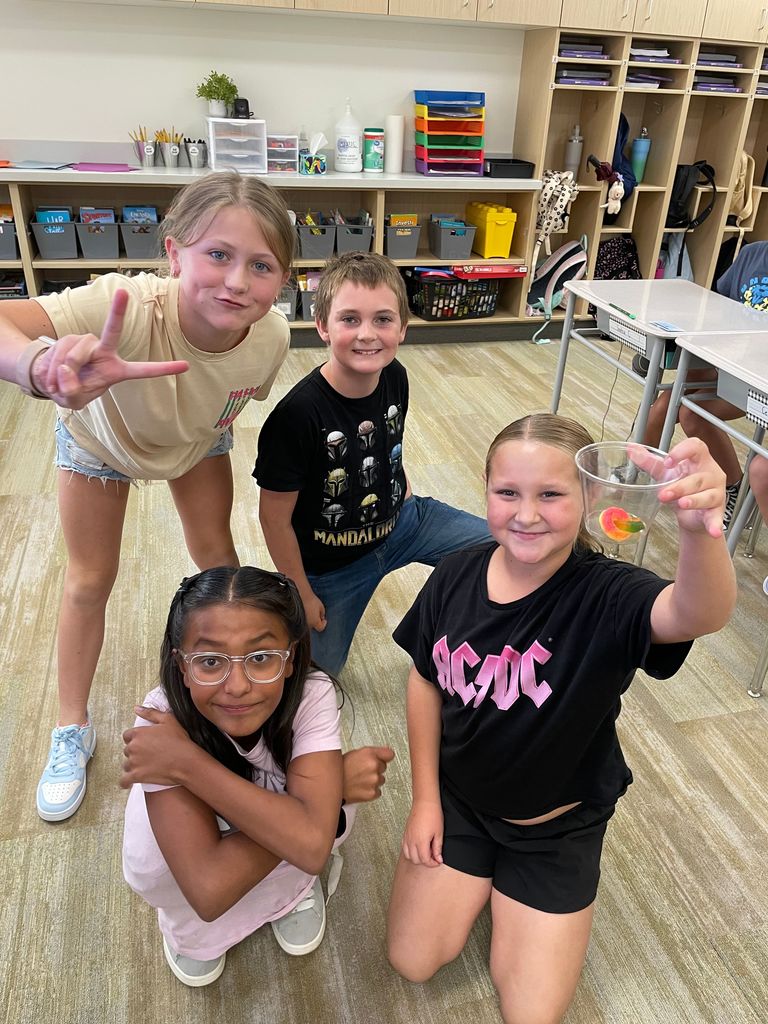 Four fifth grade students smile and pose together in a classroom. One girl kneels in front with glasses, another makes a peace sign, a boy kneels in the middle, and another girl holds up a clear cup with a gummy worm inside.