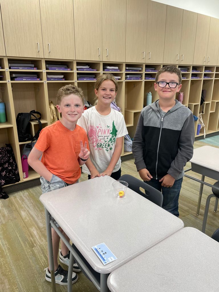 Three fifth grade students stand together by a classroom desk. The boy on the left wears an orange shirt and flashes a peace sign, the girl in the middle wears a white shirt with pink writing, and the boy on the right wears glasses and a gray hoodie. A clear cup with a gummy worm is on the desk in front of them.