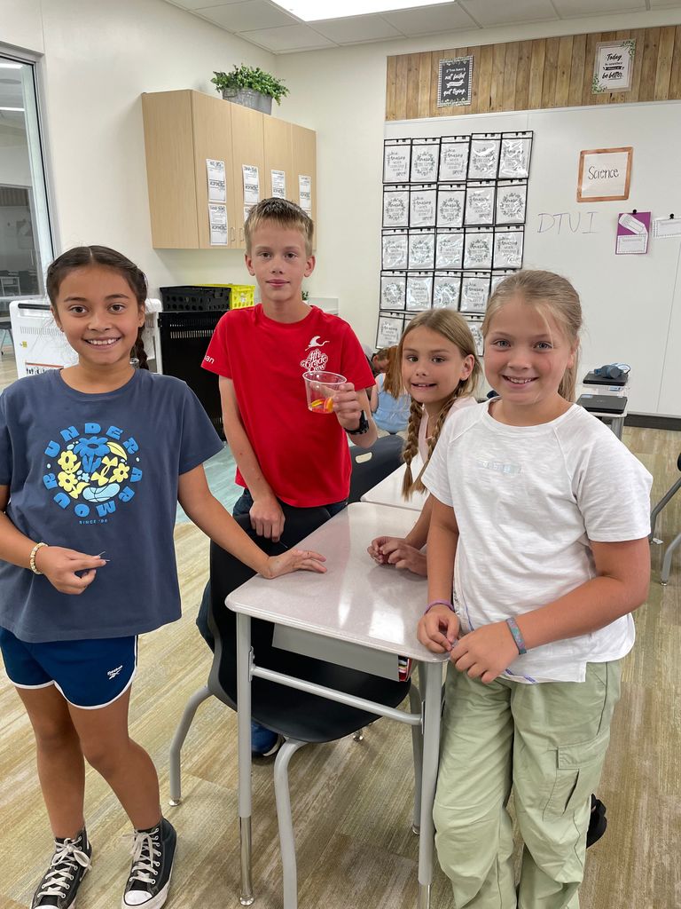 Four fifth grade students stand smiling around a classroom desk. A girl in a navy shirt stands on the left, a boy in a red shirt holds a clear cup with a gummy worm, and two girls in white shirts stand on the right.