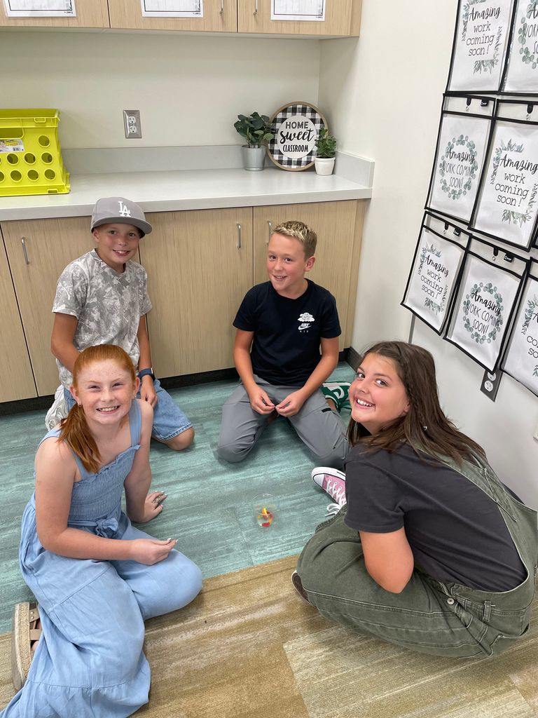 Four children sit on the classroom floor in a group, smiling and engaged. A small plastic container with candy corn is placed in the center. The background shows wooden cabinets, a countertop with plants, and a round sign that says "Home Sweet Classroom." A display of student work folders is visible on the right.