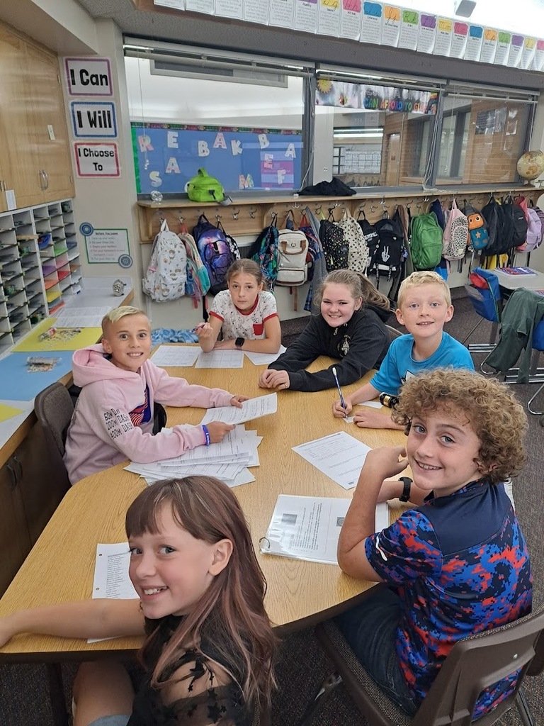 A group of smiling elementary school students sits around a table, collaborating on a classroom assignment.