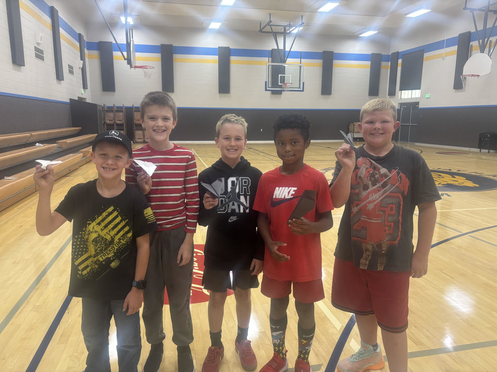There were five children in the gymnasium, each holding a paper airplane. They were standing on a wooden basketball court with yellow and blue markings.