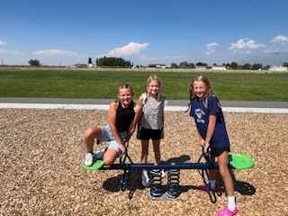 Three young girls smile while playing on a spring-based seesaw in a playground area covered with wood chips. Behind them is a wide grassy field and a clear sky with a few clouds.