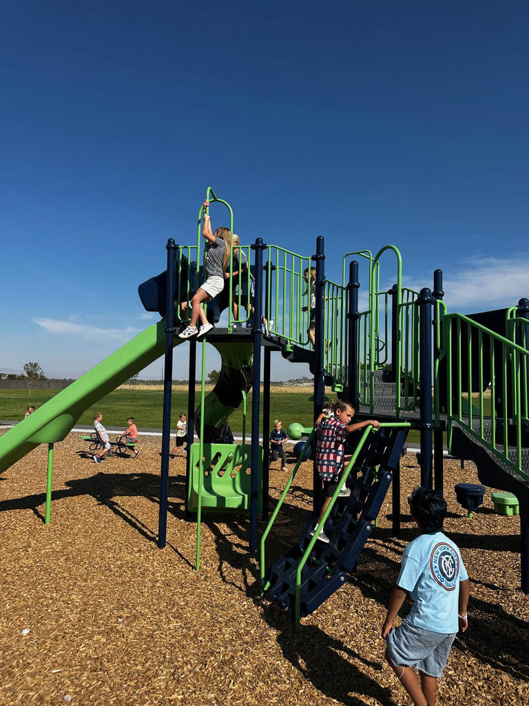 Children play actively on a large green and navy blue playground structure. Some are climbing while others are using different equipment. The sky is clear and blue, and the area is surrounded by grassy fields.