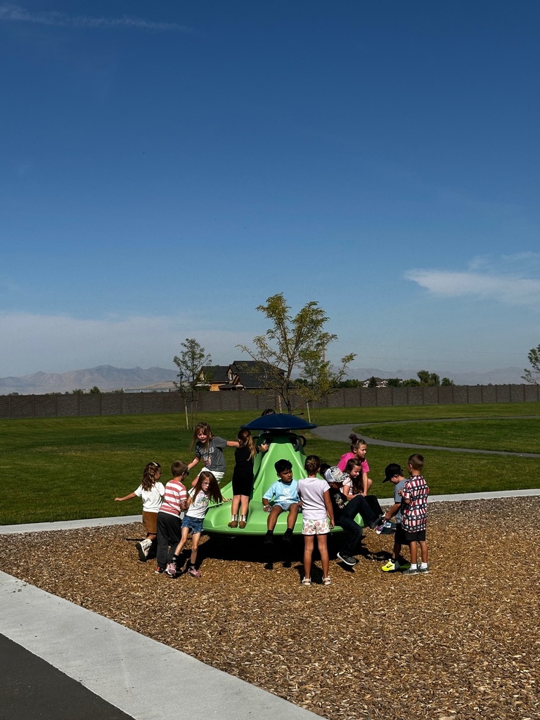 A large group of elementary students gather around and play on a green spinner structure during recess. Some are sitting on the equipment while others are running around it. The background features grassy fields, a paved path, and a blue sky.