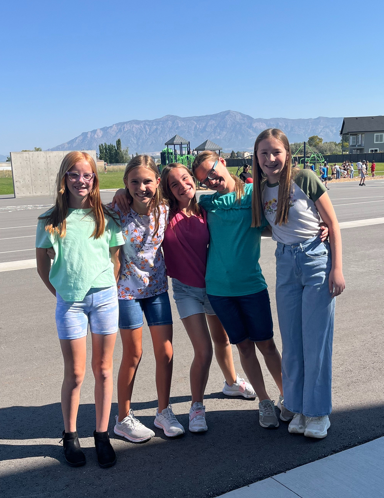 A group of five elementary school girls stand close together, smiling at the camera during recess. They’re outside on a sunny day with a scenic mountain range in the background and a playground in the distance.
