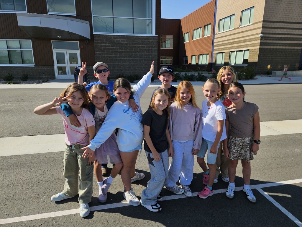 A cheerful group of ten students pose together and smile in front of the school building. Some flash peace signs while others lean in with excitement, enjoying their break on a sunny day.