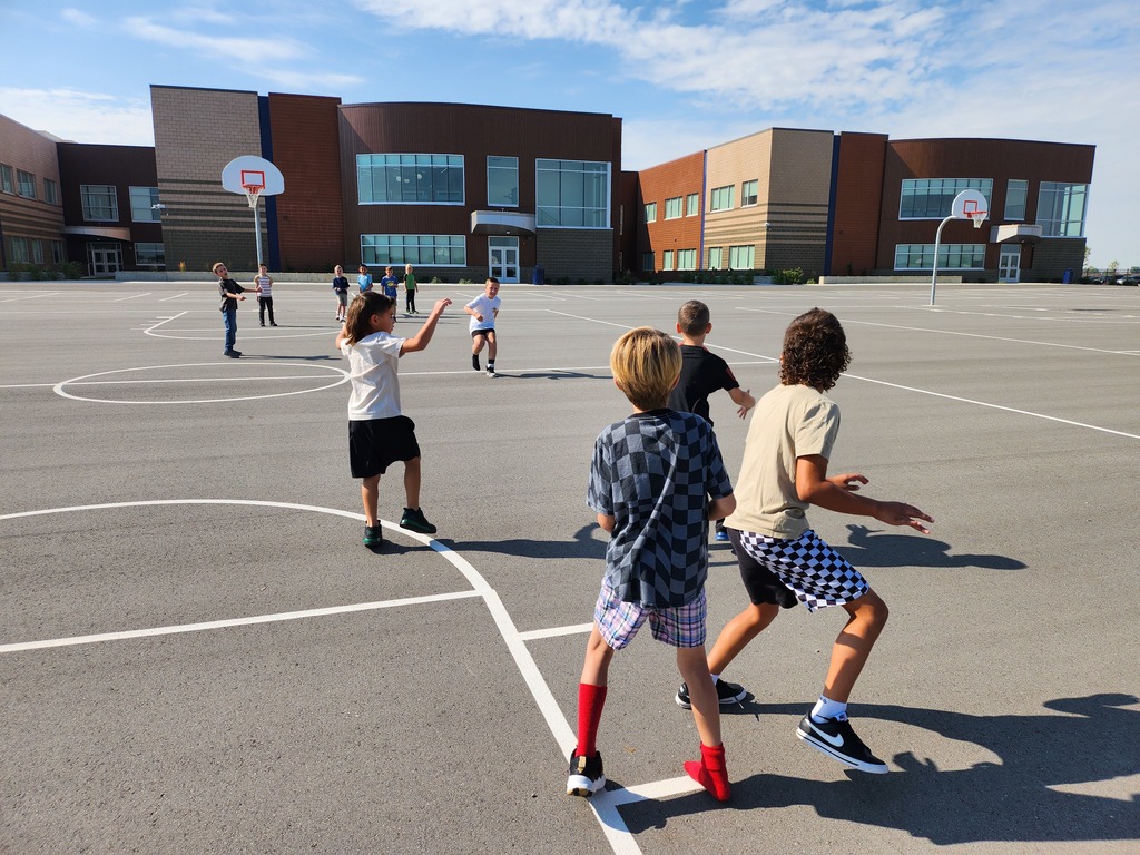 Several students are playing a basketball game on the large blacktop outside the school. Some are chasing the ball while others are positioned defensively. The school building and two basketball hoops are visible in the background.
