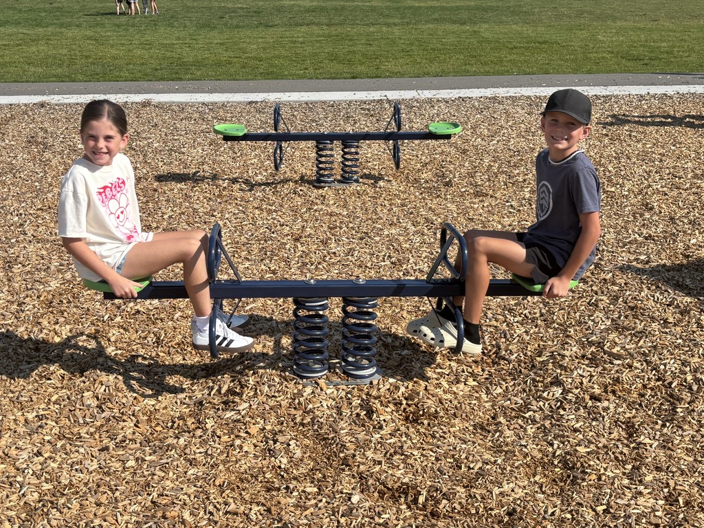 A young girl and boy sit on opposite ends of a spring-based seesaw, smiling at the camera. They’re on a wood chip-covered playground area with more green space and children in the distance.