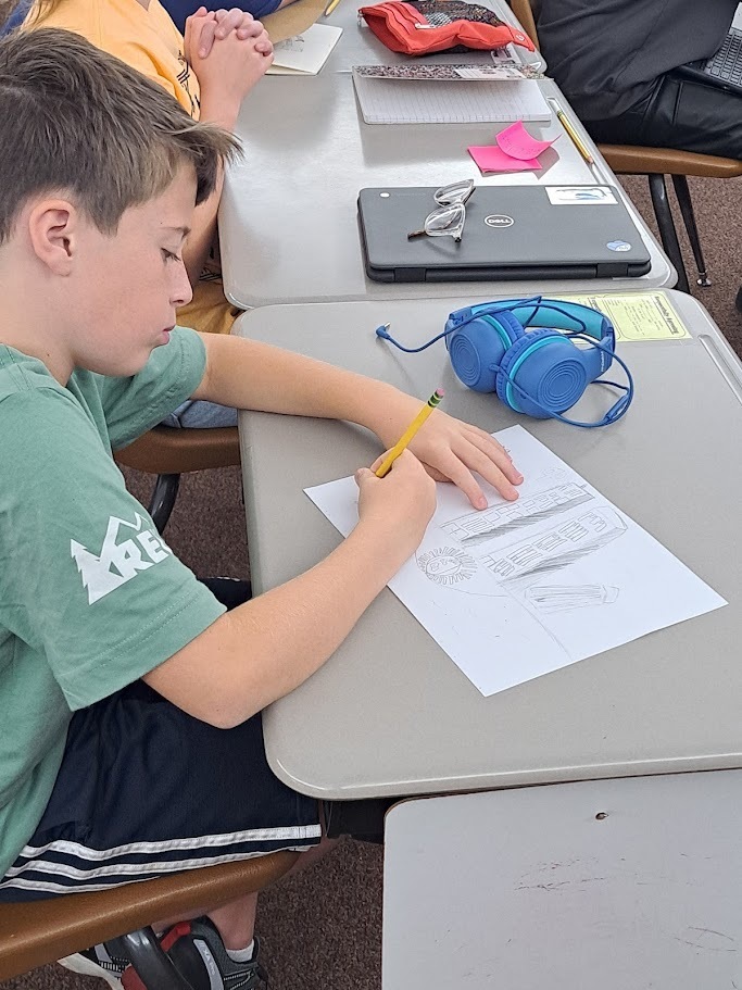 A young student sits at a classroom desk, focused on drawing a detailed picture of two buildings with a pencil. A laptop and blue headphones are on the desk next to him.