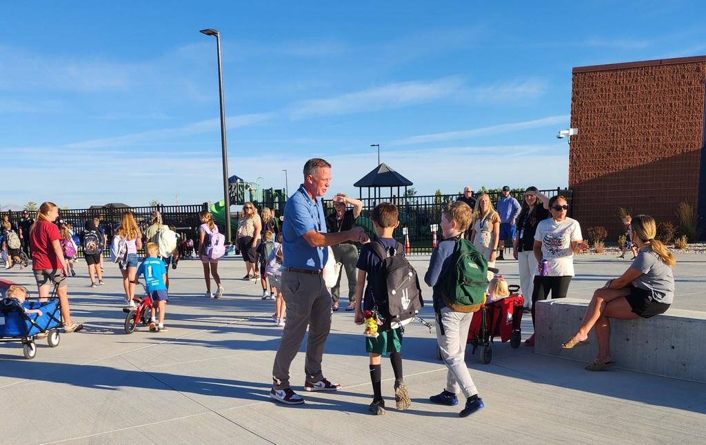 The school principal, Mr. Skeen, greets students arriving at school under a clear blue sky. Children with backpacks and parents fill the busy concrete courtyard.